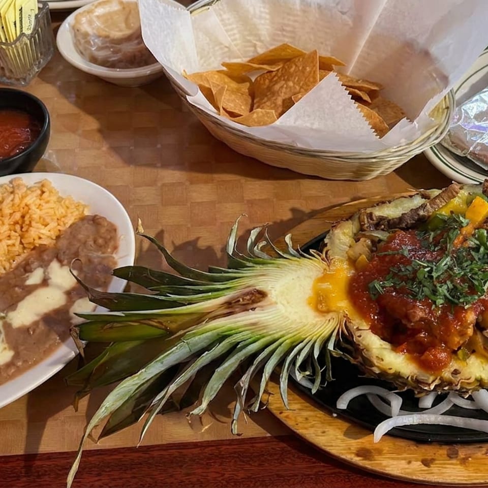 Mexican restaurant meal featuring pineapple-topped grilled meat, rice, beans, guacamole, sour cream, salsa, and tortilla chips.