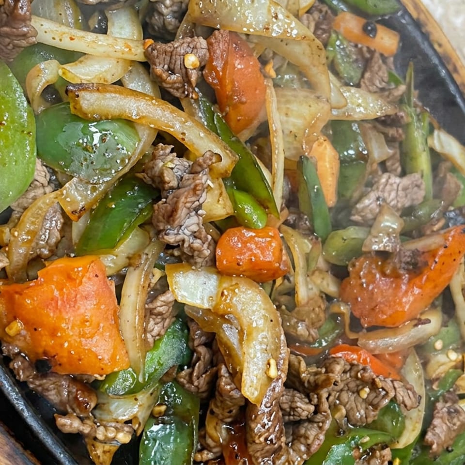 Sizzling beef strips with green peppers, onions, carrots, and tomatoes served on a black skillet.