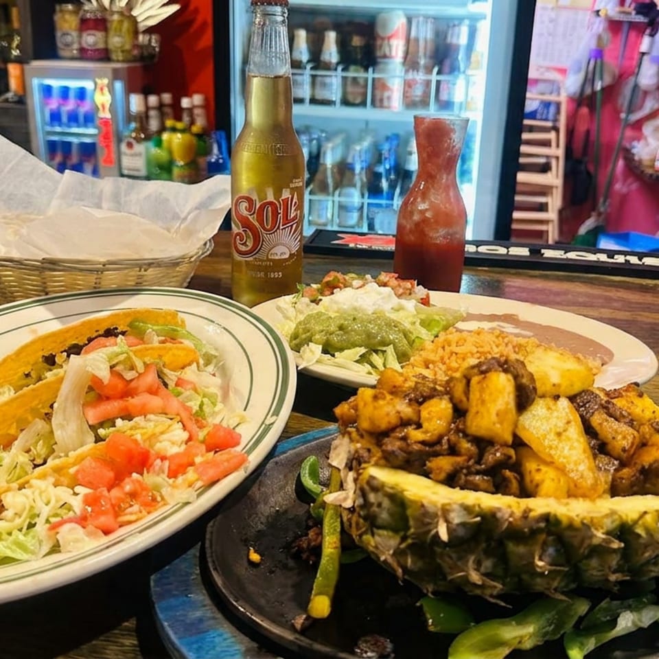 Mexican restaurant counter displaying tacos, a pineapple stuffed with grilled meat, rice, guacamole, salsa, and a Sol beer.